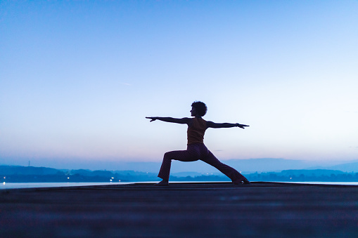 Mature woman doing yoga on wooden pier above the beautiful lake at dusk. She is dedicated to healthy lifestyle