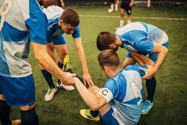 Soccer players helping their injured teammate Group of men, male soccer player lying on grass injured in soccer match, his teammates are helping him. soccer team captain stock pictures, royalty-free photos & images