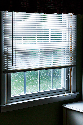 Looking out through the opaque window from a dark bedroom in a suburban residential house at the window screen drenched from a springtime extreme weather downpour rain storm outside.