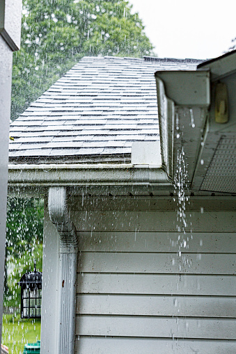 Drenching downpour rain storm water is overflowing off the tile shingle roof - dripping, spraying and splashing on over the eaves trough aluminum roof gutter system on a suburban residential colonial style house near Rochester, New York State, USA during a torrential May late spring thunderstorm.