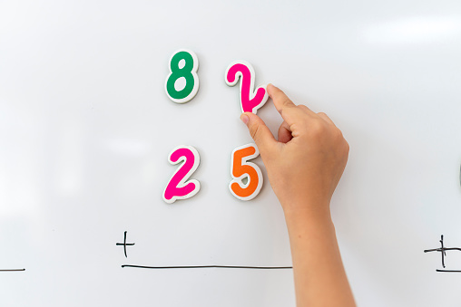 Primary school student in uniform solving math problem with magnetic numbers on whiteboard.