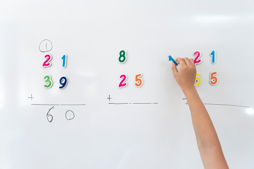 Primary school student in uniform solving math problem with magnetic numbers on whiteboard.