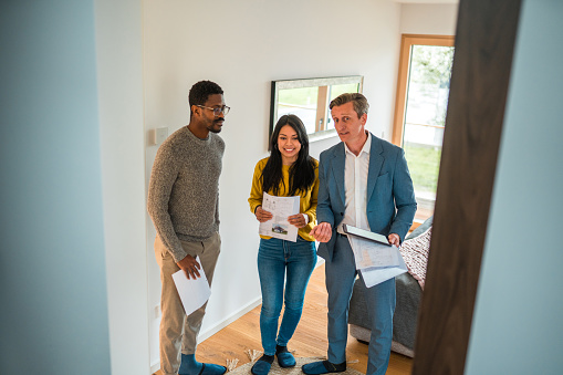 Young Hispanic woman and African American man in a meeting with a caucasian salesman and real estate agent. They are walking indoors and looking at the rooms in a house for sale.
