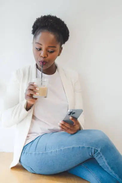Photo of beautiful afro woman using mobile phone while sitting at cafe, drinking an iced-coffee. Social networking. Social media. Modern lifestyle.