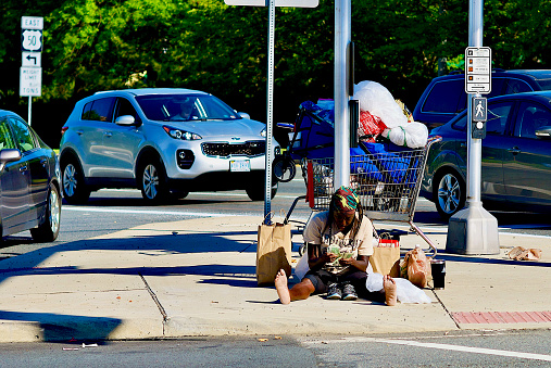Fairfax, Virginia, USA - June 8, 2020: A homeless person surrounded by their possessions counts dollar bills while sitting on the ground at a major intersection in the City of Fairfax.