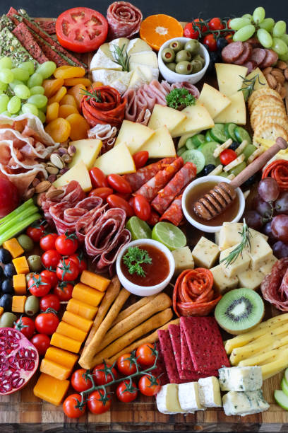 istock Full frame image charcuterie board ingredients, crackers, ham, prosciutto and salami roses, cucumber, lime half, cherry tomatoes, Brie, kiwi, Cheddar, red grapes, ramekin of honey and honey dipper, black background, elevated view 1440141616
