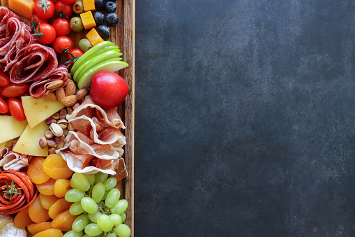 Stock photo showing close-up, elevated view of wooden charcuterie board covered with prepared sliced and chopped ingredients including breadsticks, ham, prosciutto and salami roses, stuffed olives, apple slices, vine tomatoes, Brie, Red Leicester, blueberries, Cheddar, nuts, apricots, grapes and a ramekin of chutney.