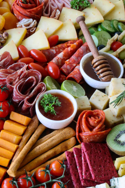istock Full frame image charcuterie board ingredients, crackers, ham, prosciutto and salami roses, cucumber, lime half, cherry tomatoes, Brie, kiwi, Cheddar, red grapes, ramekin of honey and honey dipper, elevated view, focus on foreground 1440141627