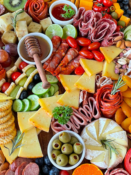 istock Full frame image charcuterie board ingredients, crackers, ham, prosciutto and salami roses, cucumber, lime half, cherry tomatoes, Brie, kiwi, Cheddar, red grapes, ramekin of honey and honey dipper, elevated view 1440141644
