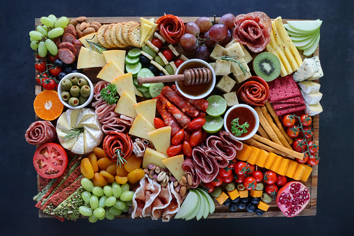 Stock photo showing close-up, elevated view of wooden charcuterie board covered with prepared sliced and chopped ingredients including crackers, ham, prosciutto and salami roses, cucumber, lime half, cherry tomatoes, Brie, kiwi, Cheddar, red grapes, ramekin of honey and honey dipper.