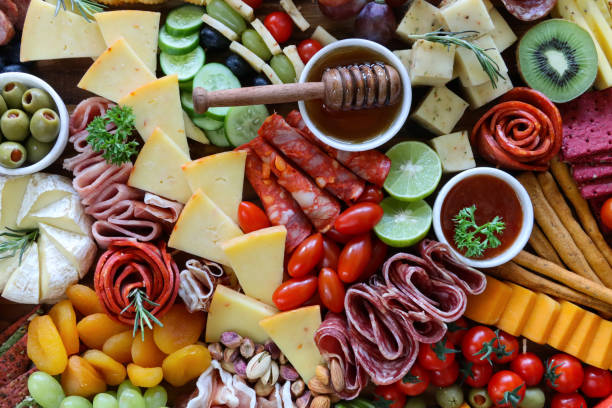 istock Full frame image charcuterie board ingredients, crackers, ham, prosciutto and salami roses, cucumber, lime half, cherry tomatoes, Brie, kiwi, Cheddar, red grapes, ramekin of honey and honey dipper, elevated view 1440141650