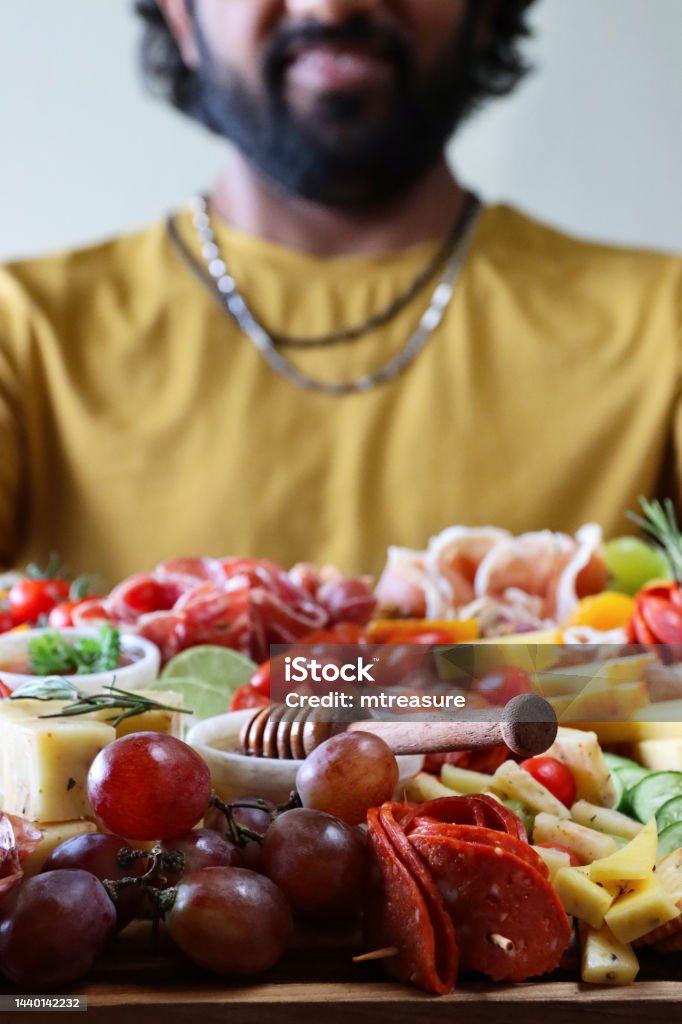 Close-up image of unrecognisable man holding charcuterie board with ramekins of honey and chutney, rows of crackers, ham and salami roses, breadsticks, kiwi, vines tomatoes, soft and hard cheeses, stuffed olives, blueberries, focus on foreground Stock photo showing close-up, view of wooden charcuterie board covered with prepared sliced and chopped ingredients including ramekins of honey and chutney, rows of crackers, ham and salami roses, breadsticks, kiwi, vines tomatoes, soft and hard cheeses, stuffed olives and blueberries displayed against blurred green garden background. 30-34 Years Stock Photo