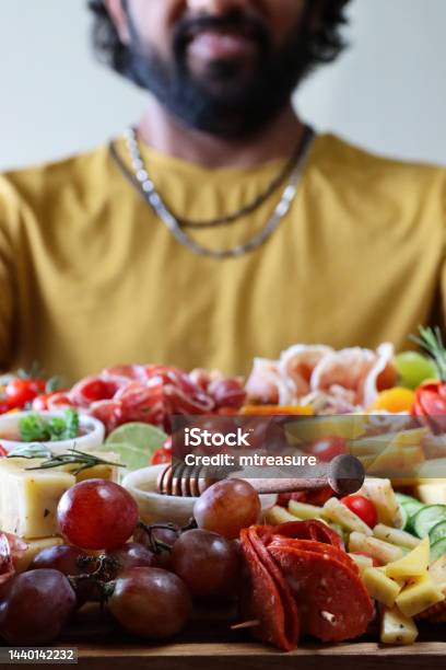 Closeup Image Of Unrecognisable Man Holding Charcuterie Board With Ramekins Of Honey And Chutney Rows Of Crackers Ham And Salami Roses Breadsticks Kiwi Vines Tomatoes Soft And Hard Cheeses Stuffed Olives Blueberries Focus On Foreground Stock Photo - Download Image Now