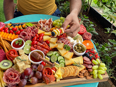 Stock photo showing close-up, view of wooden charcuterie board covered with prepared sliced and chopped ingredients including ramekins of honey and chutney, rows of beetroot crackers, ham and salami roses, breadsticks, kiwi, vines tomatoes, soft and hard cheeses, stuffed olives and blueberries displayed on an apartment balcony table.