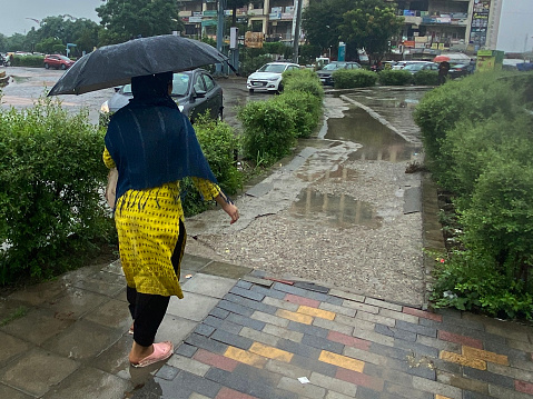 Noida, Uttar Pradesh, India - September 10, 2022: Stock photo showing rear view of an unrecognisable person walking through puddles on paved pathway whilst carrying an open umbrella as protection from downpour of rain.