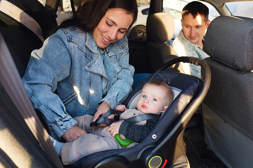 Child kid baby boy in safety seat near to mother. Dad sits on forward sitting of  car. Child transportation safety.