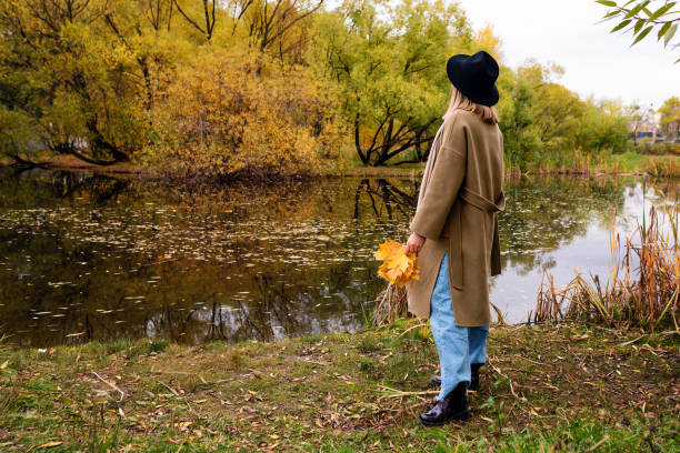 Full length unrecognizable young woman in coat, scarf and hat in autumn season. Girl holds yellow fallen maple leafs in hands and looks at pond or lake in park.