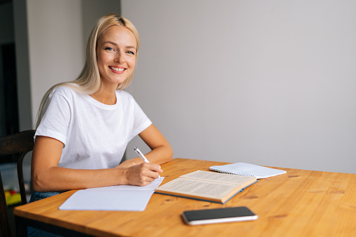 Portrait of cheerful blonde female student writing notes or essay, studying, reading textbook, sitting at table with books, smiling looking at camera, preparing for university exams, doing homework.