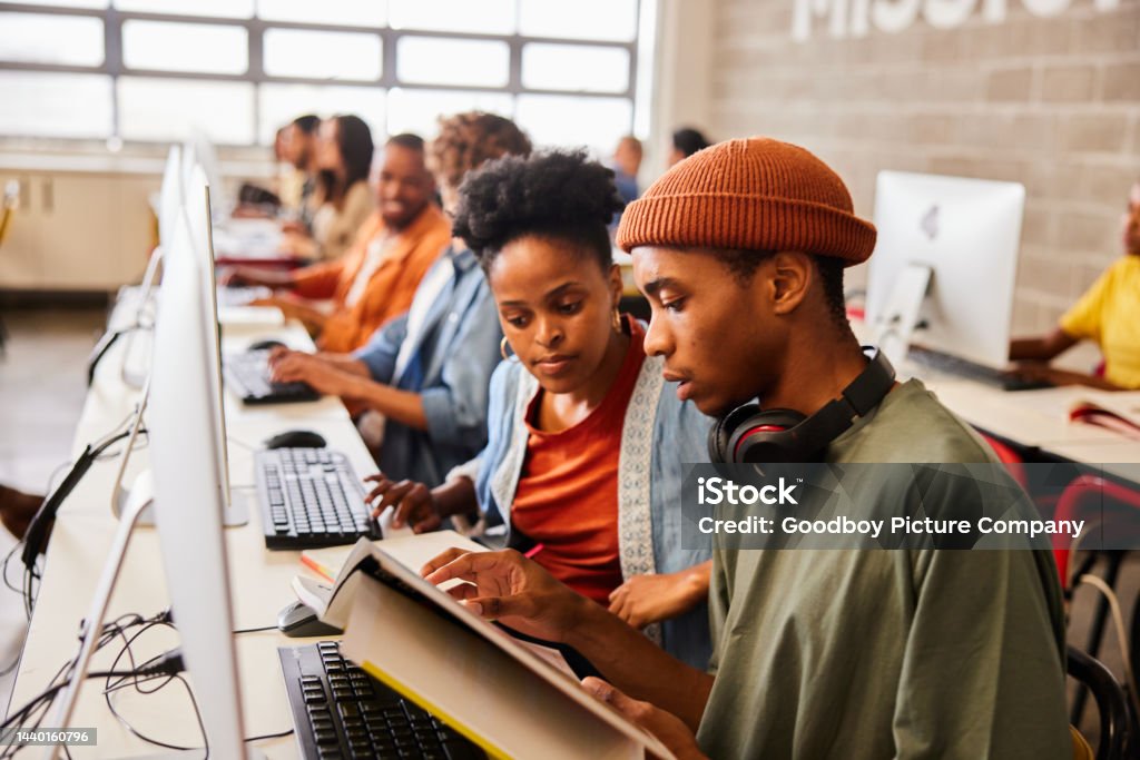 Two college students reading a textbook during computer class Two young college students reading through a textbook while sitting together at a table in computer class University Student Stock Photo