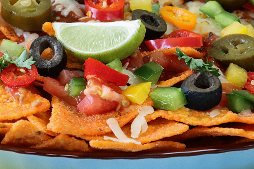 Stock photo showing close-up, elevated view of homemade loaded nachos recipe. This snack contains fresh, crunchy tortilla chips covered in melted cheese with cherry tomatoes, black olives and Jalapeno peppers on a blue table with a blue plate, served with lime wedge, salsa, sauces and dips.