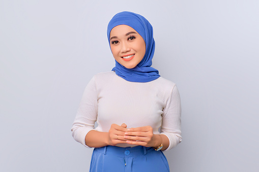 Smiling young beautiful Asian Muslim woman holding hands together and feeling confident isolated over white background