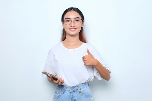 Smiling young Asian girl in white t-shirt using smartphone and showing thumb up isolated on white background