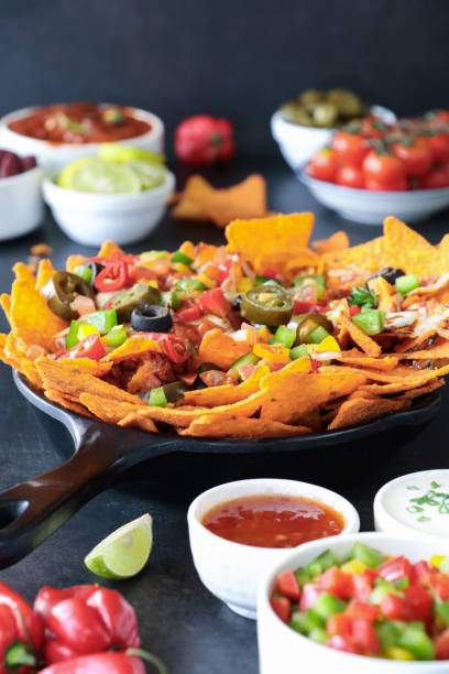 istock Full frame image of homemade, loaded nachos covered in melted mozzarella cheese, cherry tomatoes, black olives, Jalapeno peppers, elevated view, focus on foreground, served with sauces and dips 1440166375