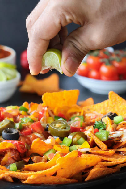 istock Full frame image of hand squeezing lime wedge over homemade, loaded nachos covered in melted mozzarella cheese, cherry tomatoes, black olives, Jalapeno peppers, elevated view, focus on foreground, served with sauces and dips 1440166380