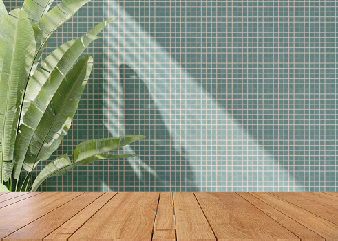 Wooden tabletop or countertop in modern turquoise mosaic wall with dappled sunlight from window and tropical banana tree at home for household and personal product display
