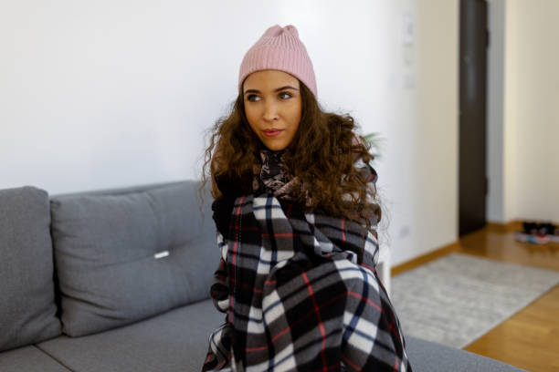 A woman in warm clothing is shivering and wearing a blanket and sitting at home freezing.