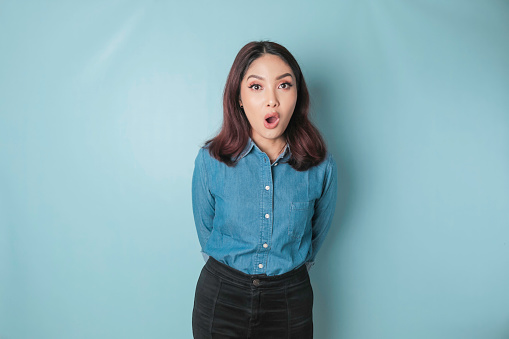 A portrait of a shocked Asian woman wearing a blue shirt, isolated by a blue background