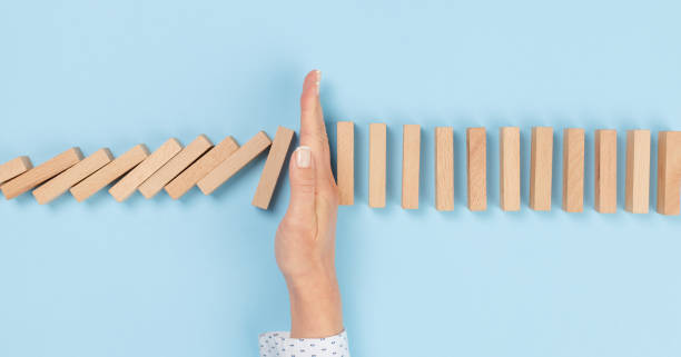 Woman hand stopping falling wooden dominoes effect on blue solid ground stock photo