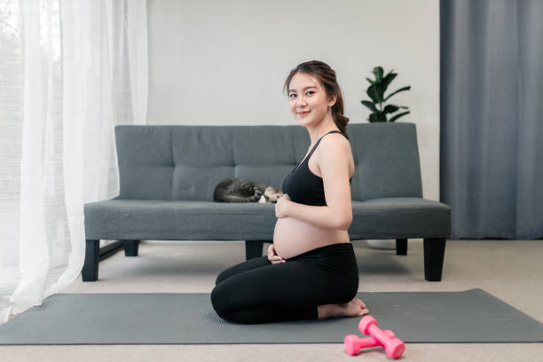 Pretty pregnant asian woman is sitting with a smile on her face while looking at the camera and her hands rubbing her pregnant tummy. stock photo
