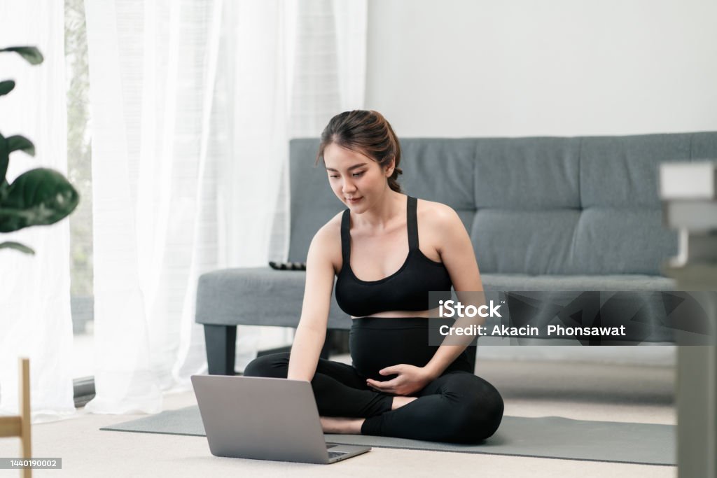 Pregnant asian woman is using her computer laptop for class online yoga to keep her prenatal healthy in the living room. Pregnant asian woman is using her computer laptop for class online yoga to keep her prenatal healthy in the living room. Concept healthy pregnancy. 20-29 Years Stock Photo