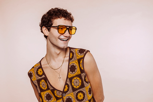 Studio portrait of a happy smiling young homosexual gay man laughing, isolated on beige background. Wears yellow stylish sunglasses and top, looks away. Copy space.