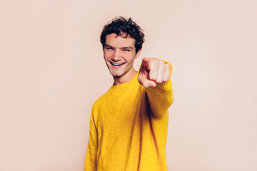 Young cheerful boy point finger at you, positive mood. Man with curly hair wears yellow sweater. Studio portrait isolated over beige background.