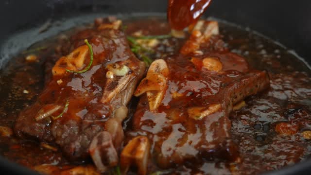 Spoon pours sauce over steak beef ribs with garlic in pan on stove. Cooking meat