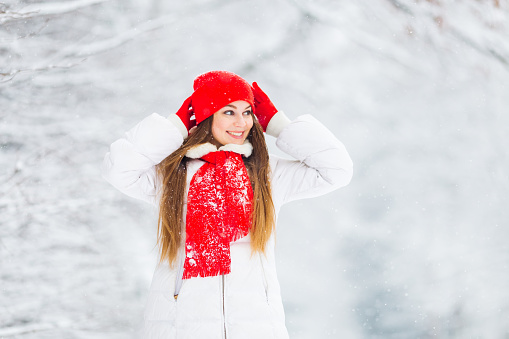The girl smiles and holds her hands in her gloves near the red hat and looks distantly on against the background of the snowy trees.