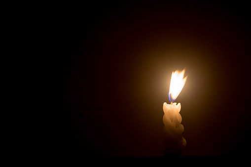 Blurred photo of single burning candle flame or light is glowing on a spiral white candle on black or dark background on table in church for Christmas, funeral or memorial service.