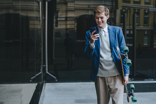 Smiling handsome young man businessman wearing suit messaging using cellphone holding longboard