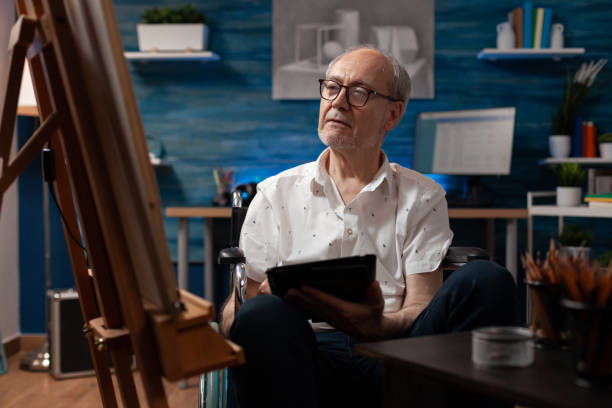 uomo anziano seduto in sedia a rotelle tenendo il computer digitale del tablet che guarda il disegno dello schizzo del vaso - romania men artist portrait foto e immagini stock