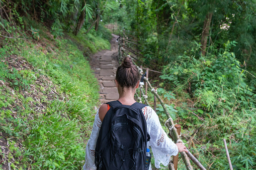 Young woman walking and leaning a hand on the railing of a catwalk of sacks placed on the ground in the middle of the Asian jungle.