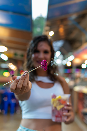 Vertical image of a woman showing a piece of dragon fruit that she has pricked on a wooden stick while holding with the other hand a plastic cup filled with Asian tropical fruit from the street market
