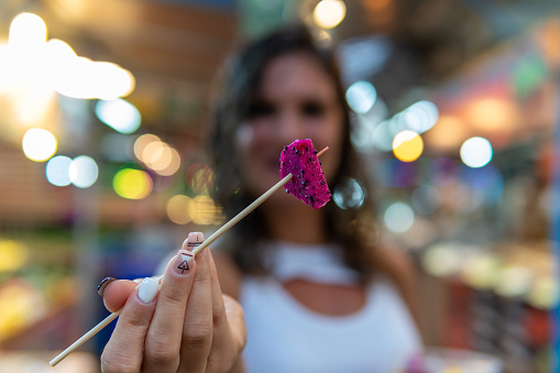 Woman showing a piece of dragon fruit that she has pricked on a wooden stick while holding with the other hand a plastic cup filled with Asian tropical fruit bought in the street market.