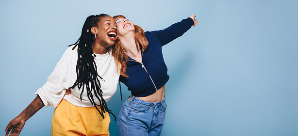 Two happy best friends laughing and having a good time while embracing each other in a studio. Two cheerful female friends making fun memories while standing against a blue background.