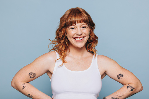 Portrait of a happy young woman with arm tattoos standing in a studio. Cheerful young woman with ginger hair standing against a blue background in a white tank top.