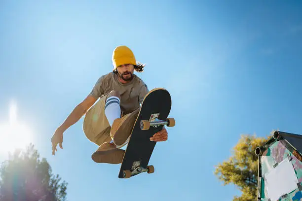 A young adult dexterous skater is jumping in midair over the camera holding to his board.
