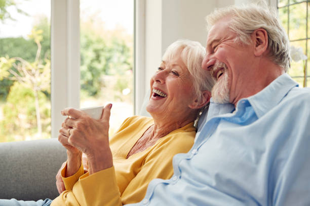 Coppia anziana in pensione che si siede sul divano a casa bevendo caffè e guardando la TV insieme - foto stock