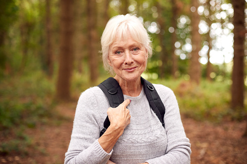 Portrait Of Active Retired Senior Woman Walking In Woodland Countryside Wearing Backpack