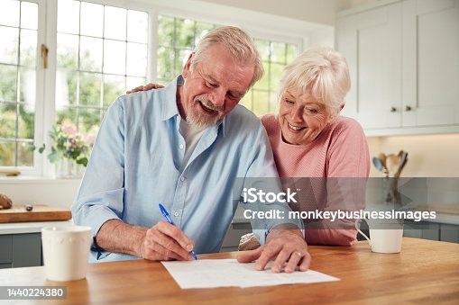 istock Retired Senior Couple Sitting In Kitchen At Home Signing Financial Document 1440224380